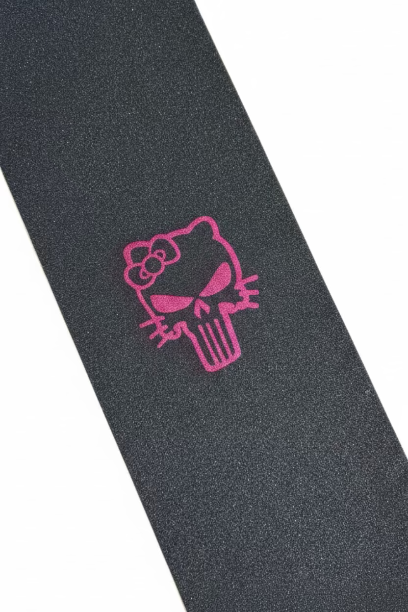 A close-up of a skateboard grip tape featuring a pink Hello Kitty Punisher skull design on a dark gray textured background, shown diagonally with cardboard boxes in the background.