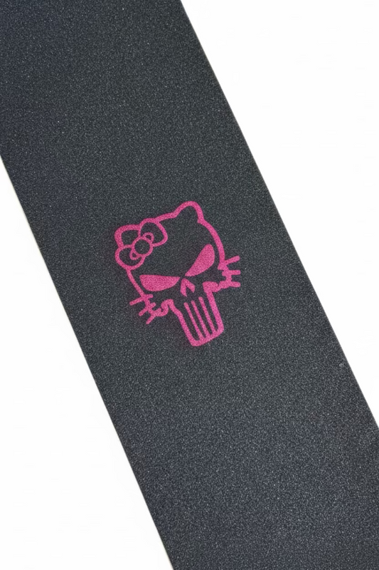 A close-up of a skateboard grip tape featuring a pink Hello Kitty Punisher skull design on a dark gray textured background, shown diagonally with cardboard boxes in the background.